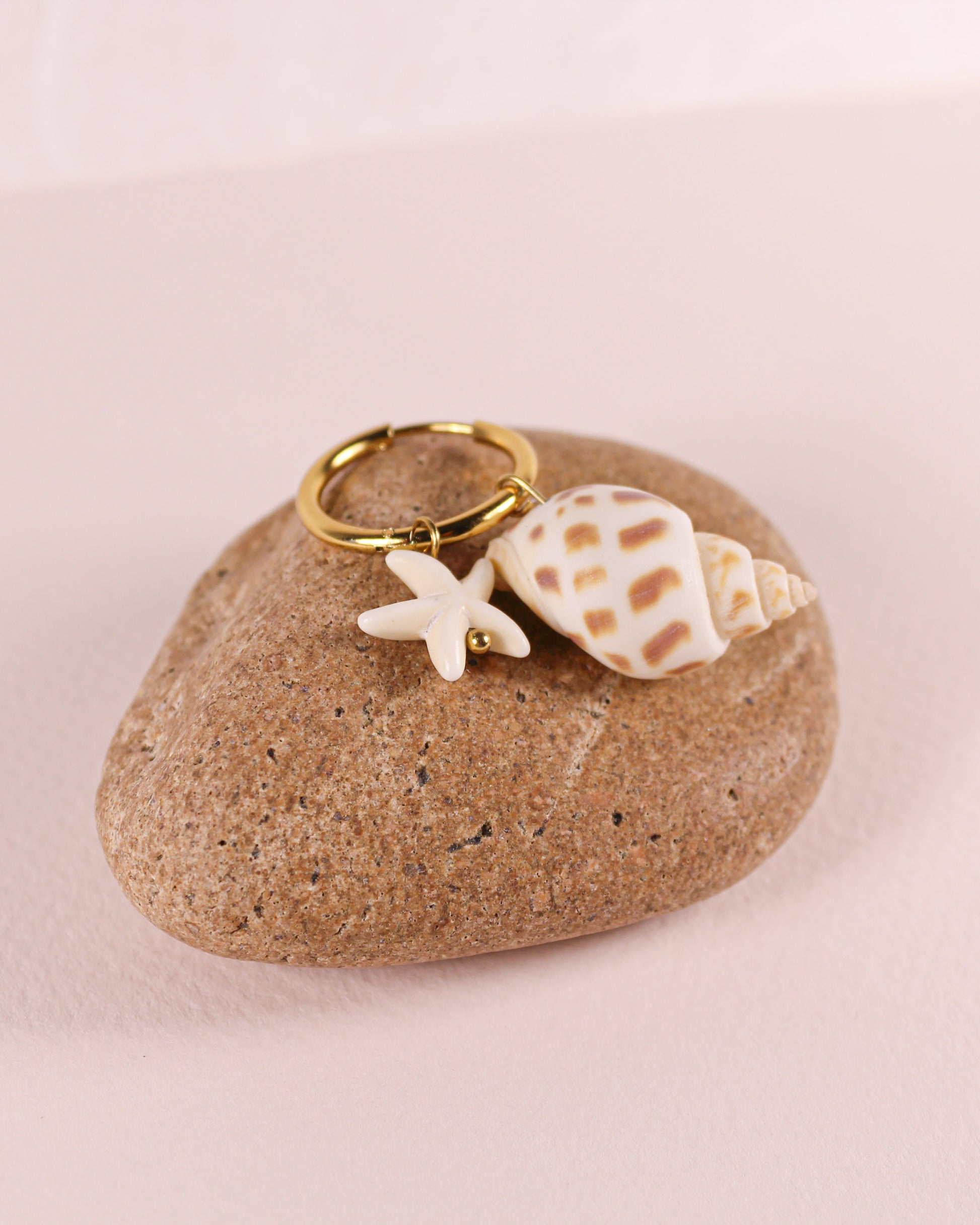 Gold earrings with starfish and shell charm on a rock against a pink background