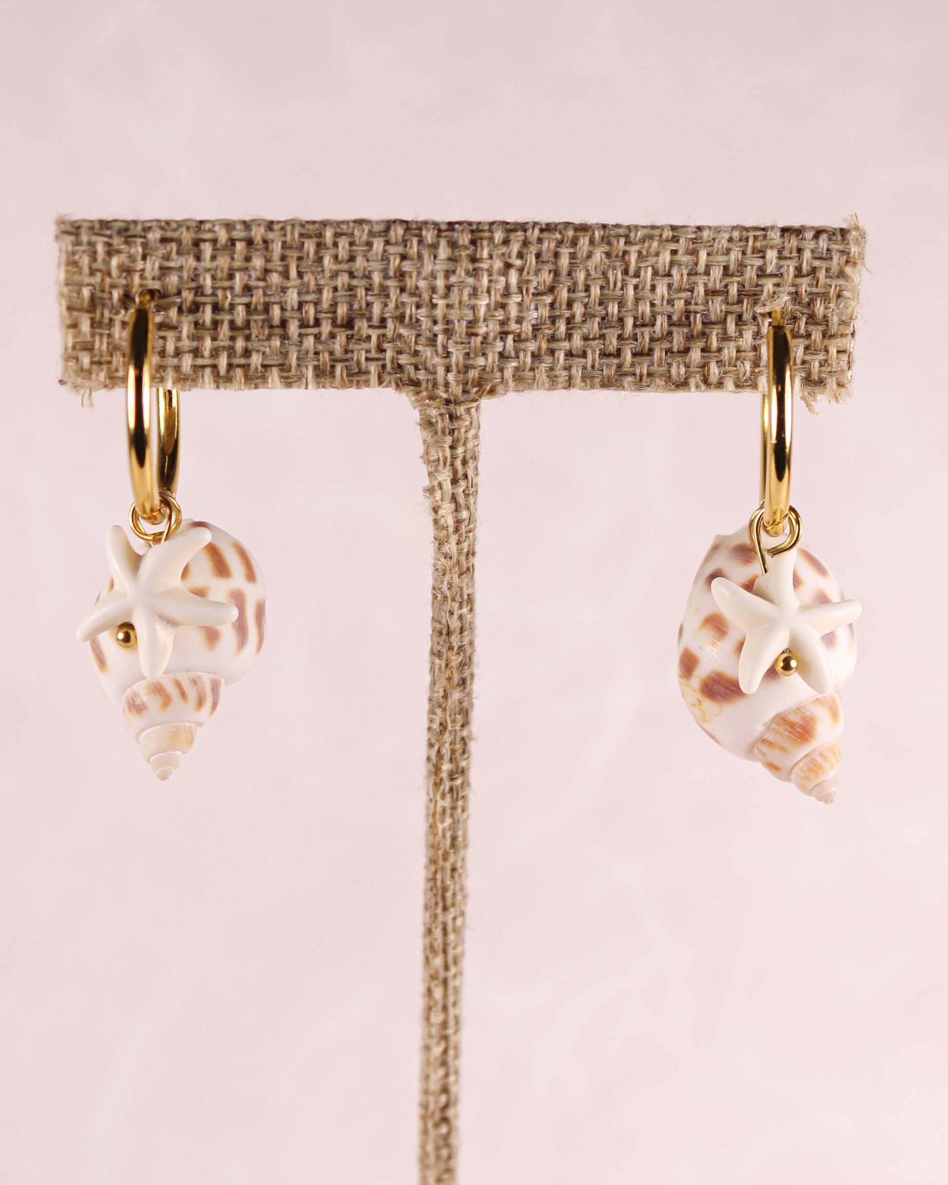 Shell and starfish earrings on a beige earring stand against a pink background