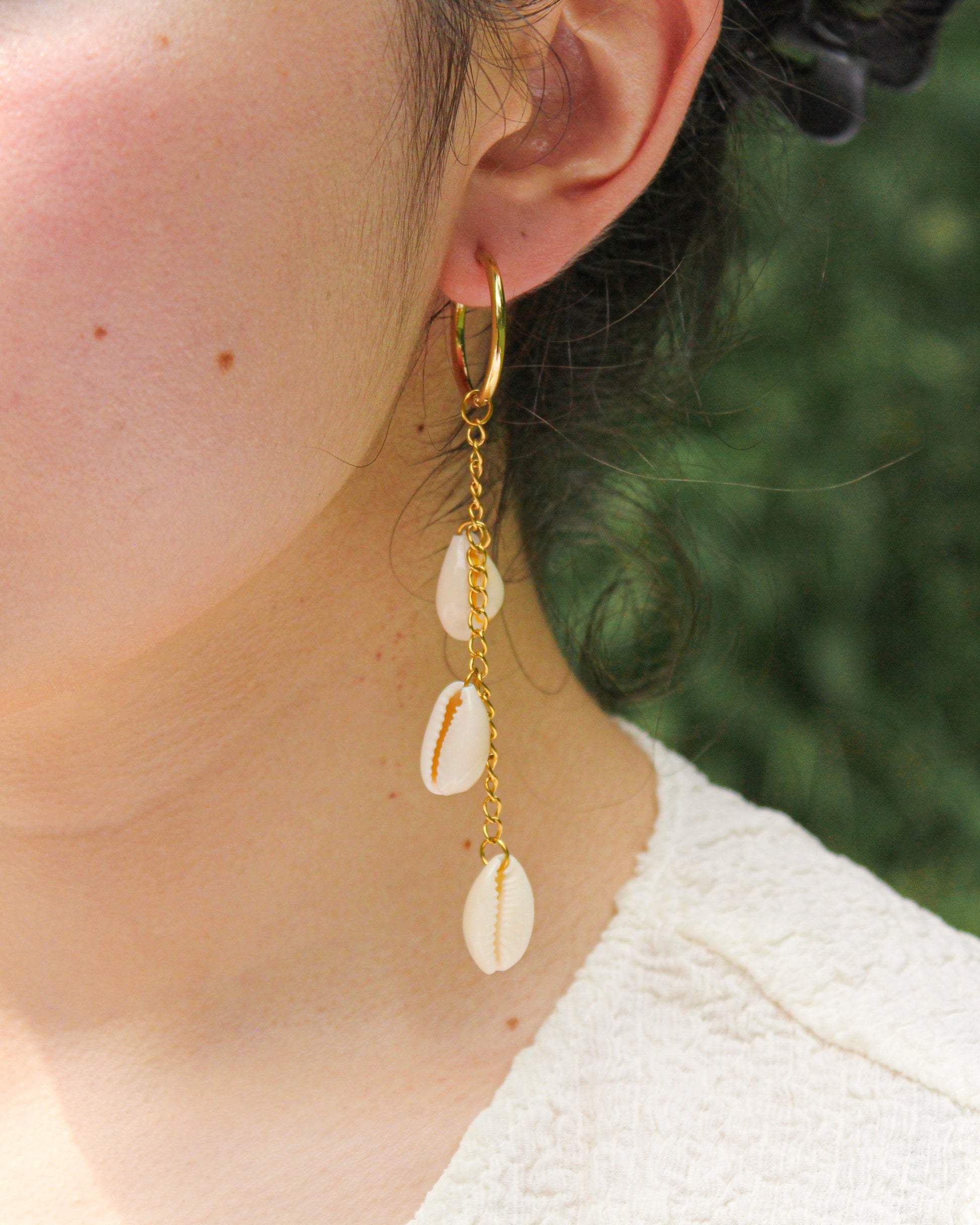 Close-up of a person wearing gold hoop earrings with shell drops against a blurred green background