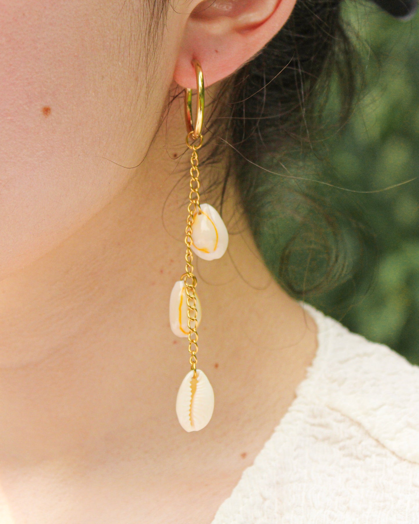 Gold hoop earring with shell charms worn by a person against a blurred green background